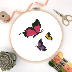 May include: A cross-stitch embroidery hoop featuring three butterflies. The largest butterfly is pink and black, the middle butterfly is purple and black, and the smallest butterfly is yellow and black.