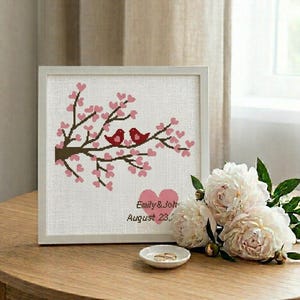 Op de afbeelding: Een ingelijst kruissteekkunstwerk met twee rode vogels op een bloeiende tak met roze bloesems. Het kunstwerk bevat de namen "Emily & John" en de datum "August 23," in een witte lijst, met een klein schaaltje met twee ringen.