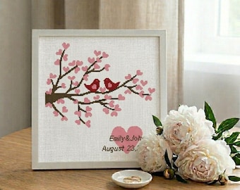 May include: A framed cross-stitch artwork featuring two red birds perched on a flowering branch with pink blossoms. The artwork includes the names "Emily & John" and the date "August 23," in a white frame, with a small dish holding two rings.