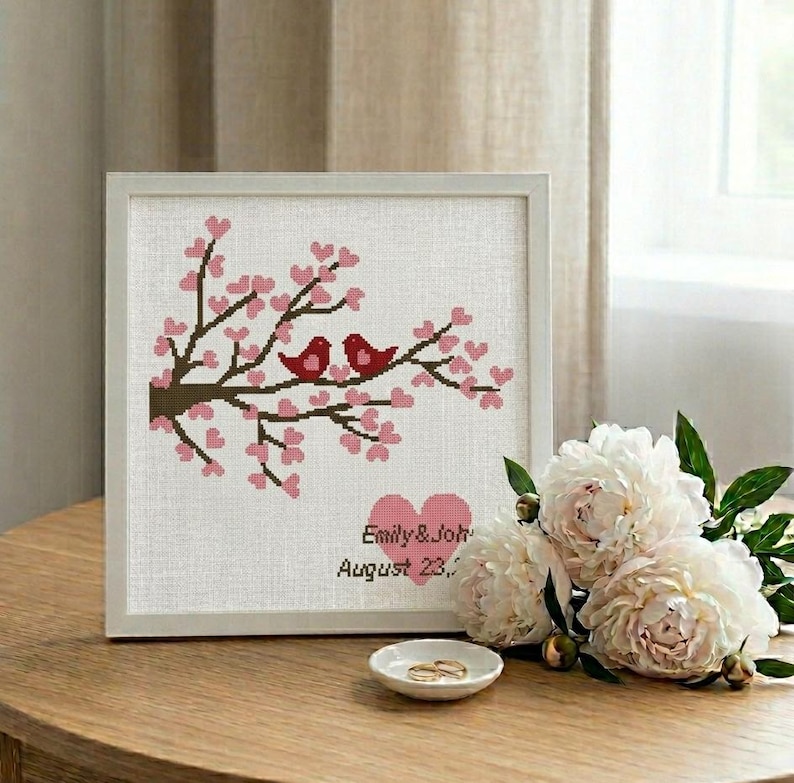Puede incluir: Una obra de arte en punto de cruz enmarcada con dos p&aacute;jaros rojos posados en una rama floreciente con flores rosas. La obra incluye los nombres "Emily & John" y la fecha "August 23," en un marco blanco, con un plato peque&ntilde;o con dos anillos.