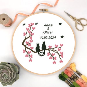 Peut inclure: Un cerceau de broderie blanc avec un motif de point de croix de deux chats noirs assis sur une branche avec des cœurs roses. Le texte "Anna & Oliver 14.02.2024" est brodé au-dessus des chats.