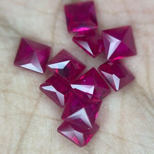 May include: A collection of square-cut, faceted ruby gemstones. The rubies are a deep, vibrant red color, with sharp angles and reflective surfaces. The gemstones are scattered, showcasing their individual brilliance and potential for jewelry design.
