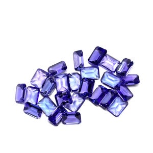 May include: A scattering of rectangular gemstones in shades of blue and purple. The gems are displayed against a white backdrop, highlighting their clarity and colour variations. The gems appear to be precisely cut, reflecting light.
