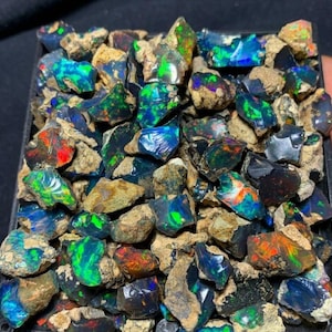 May include: A collection of rough, uncut opals in a black tray. The opals display vibrant colors including blue, green, red, and orange, with a play of light. The opals are surrounded by a matrix of brown rock.