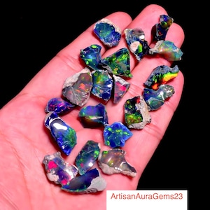 May include: A collection of rough opal gemstones, showcasing vibrant colors of blue, green, pink, and orange. The opals are held in a hand, with a black background. The text "ArtisanAuraGems23" is visible in the lower right corner.