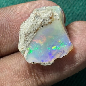 May include: A rough opal gemstone with a white base and iridescent flashes of green, blue, and yellow. The stone has a rough, textured surface with a light-colored matrix.