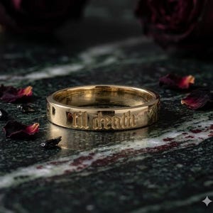 Könnte beinhalten: Ein polierter Goldring mit der eingravierten Aufschrift 'til death' in einer Schriftart im gotischen Stil. Der Ring hat eine glatte, reflektierende Oberfläche und ein flaches Profil, das die detaillierte Gravur im Metallband hervorhebt.