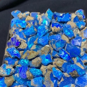 May include: A collection of raw, uncut blue opals. The opals have a vibrant, electric blue color with a translucent quality. They are mixed with rough, tan-colored matrix rock, creating a natural, earthy contrast.
