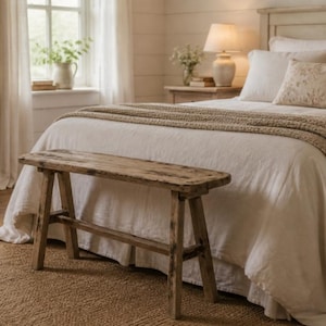 Könnte beinhalten: Eine rustikale Holzbank, etwa 120 cm lang, steht am Fuß eines Bettes mit weißer Bettwäsche und einer beigefarbenen Strickdecke. Der Raum hat eine neutrale Farbpalette, einen Jute-Teppich und eine Holzkommode. Eine Lampe und ein Fenster sind sichtbar.