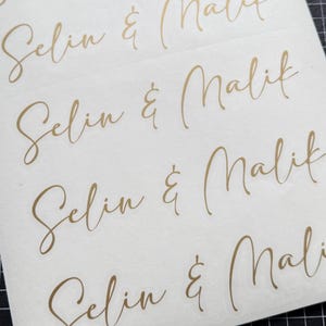 May include: Close-up of several stickers with the names "Selin & Malik" in elegant gold script. The names are repeated multiple times on a white background, with a grid pattern visible in the corner. The script is a flowing, cursive font.