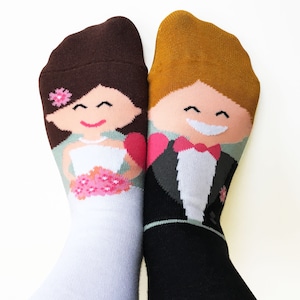 Könnte beinhalten: Ein Paar Socken mit einem Braut- und Bräutigam-Design. Die Braut trägt ein weißes Kleid mit einem rosa Blumenstrauß und einer rosa Blume im Haar. Der Bräutigam trägt einen schwarzen Smoking mit einer weißen Fliege. Die Socken sind weiß und schwarz.