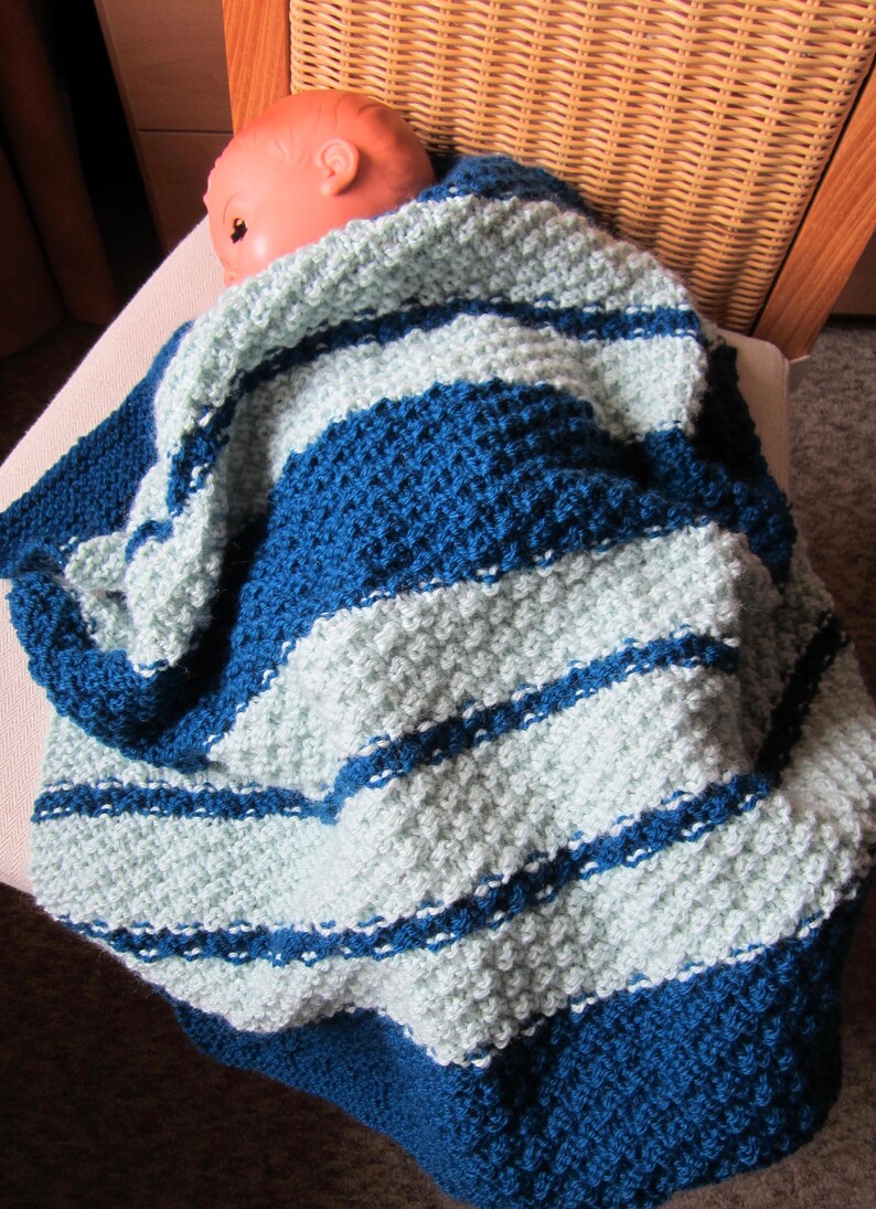 Peut inclure: Une couverture pour b&eacute;b&eacute; en tricot ray&eacute;e bleu et bleu clair. La couverture est faite avec un point textur&eacute; et a une sensation douce et confortable.