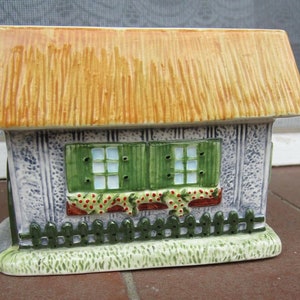 Könnte beinhalten: Eine Keramik-Häuschen-Figur mit einem Strohdach, grünen Fensterläden und einem weißen Lattenzaun. Das Häuschen hat einen roten Backsteinkamin und einen grünen Rasen.