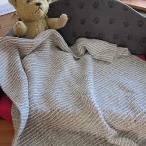 Könnte beinhalten: Eine graue und weiße gestreifte Strickdecke mit einem Teddybären darauf. Die Decke liegt auf einem grauen Hundebett mit Pfotenabdrücken.