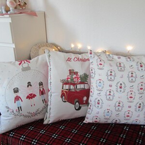 Könnte beinhalten: Drei dekorative Kissen zum Thema Weihnachten auf einer rot karierten Decke. Die Kissen zeigen festliche Designs, darunter ein roter Van mit Geschenken, Ornamenten und Nussknackerfiguren. Der Text "At Christmas" ist sichtbar.