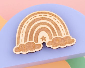 May include: Wooden rainbow and cloud cutouts. The rainbow has a star in the center and is made of light brown wood. The clouds are also made of light brown wood. The background is a mix of pastel colors, including pink, blue, and orange.