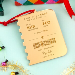May include: A gold-colored luggage tag with a red string. The tag is shaped like a ticket and has the text "PACK YOUR BAGS WE ARE GOING..." and "HAPPY CHRISTMAS Rachel LOVE, PETER X". It also includes flight details.