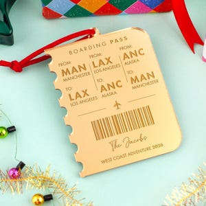 May include: A gold-colored boarding pass ornament with a red ribbon. The ornament has the text "BOARDING PASS" and details for flights from Manchester to Los Angeles and Anchorage. The text "The Jacobs WEST COAST ADVENTURE 2026" is also visible.