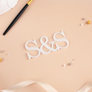 May include: A white wooden "S&S" monogram sits on a peach background, with a gold and black fork, ribbon, and decorative beads. The image is styled for a wedding or event. The overall aesthetic is soft and elegant, with a focus on detail.