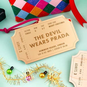 以下が含まれることがあります： 「THE DEVIL WEARS PRADA」とイベント詳細が記載された金色のチケット。ピンク色の紐で結ばれ、クリスマスの装飾に囲まれています。イベントはロンドンで開催されます。
