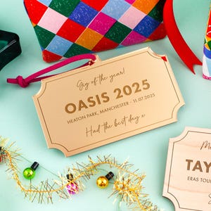 Puede incluir: Una etiqueta met&aacute;lica dorada con el texto "Gig of the year! OASIS 2025 HEATON PARK, MANCHESTER - 11.07.2025 Had the best day x". La etiqueta est&aacute; sujeta a un cord&oacute;n rosa. Otras bolsas de regalo coloridas y luces decorativas est&aacute;n en el fondo.
