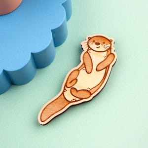 May include: A wooden otter-shaped pin with a smiling face and brown and tan coloring. The pin is lying on a light green surface. The otter has a light tan body and brown accents.