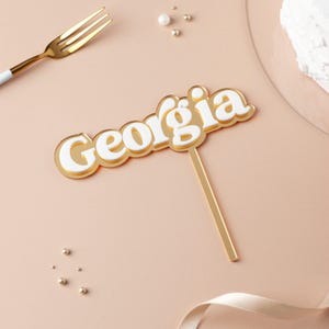 May include: Gold cake topper with the word "Georgia" in white letters. The letters are raised and have a retro style.