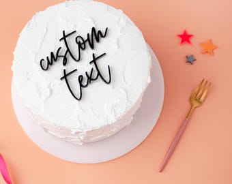 May include: A round cake covered in white frosting with a black acrylic cake topper featuring the words custom text in a cursive script. The cake is accompanied by a gold and pink fork and three small star-shaped decorations in red, orange, and gray.