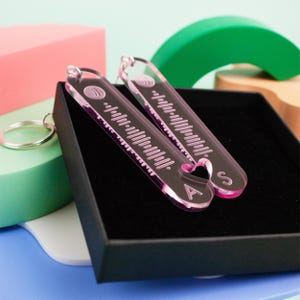 May include: Two pink acrylic keychains with a heart shape and the letters "A" and "S" on them. The keychains have a barcode-like pattern on them.