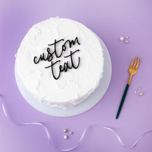 Peut inclure: Un gâteau rond blanc givré avec les mots "custom text" en écriture noire. Le gâteau est sur une assiette blanche, avec une fourchette dorée et verte. Le fond est violet, avec des perles et un ruban décoratif.