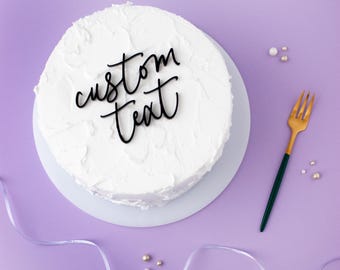 May include: A round cake covered in smooth white frosting, topped with a black script cake topper that reads custom text. A gold-toned fork with a dark green handle is positioned to the right, accompanied by small white and gold decorative pearls.