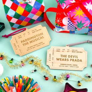 以下が含まれることがあります： 「Paddington The Musical」と「The Devil Wears Prada」のイベント詳細が記載された木製チケットオーナメント。お祝いのギフト包装とカラフルな装飾がチケットを囲んでいます。チケットはベージュで、濃い茶色の文字が書かれています。