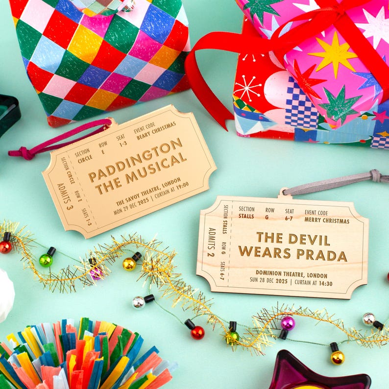 以下が含まれることがあります： 「Paddington The Musical」と「The Devil Wears Prada」のイベント詳細が記載された木製チケットオーナメント。お祝いのギフト包装とカラフルな装飾がチケットを囲んでいます。チケットはベージュで、濃い茶色の文字が書かれています。
