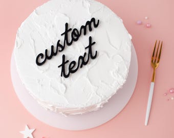 May include: A round cake covered in white textured frosting with a black script cake topper that reads custom text. A gold and white fork is positioned to the right of the cake, and small white and red star-shaped decorations are nearby.