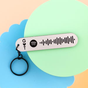 May include: A light-colored wooden keychain with a black Spotify code and a black keyring. The keychain has the letters "G" and "T" with a heart symbol. The background features a blue and green circular design.