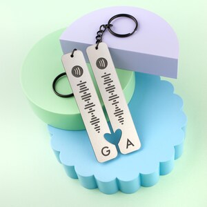May include: Two silver keychains with a music code design and a heart-shaped cutout. The keychains have the letters "G" and "A" on them. The keychains are on a blue, green, and purple background.