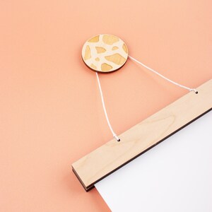 May include: A wooden hanging system with a white string and a round wooden knob with a light brown and white patterned design. The hanging system is attached to a white poster.