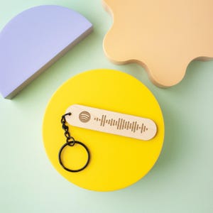 May include: A wooden keychain with a black chain and a Spotify logo and barcode engraved on it. The keychain is on a yellow surface.