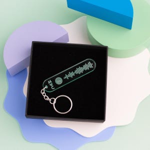 May include: A green acrylic keychain with a Spotify code and the letters "M♥C" engraved on it. The keychain is in a black box and is on a white surface.