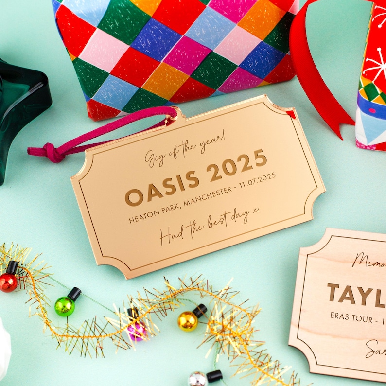 Puede incluir: Una etiqueta met&aacute;lica dorada con el texto "Gig of the year! OASIS 2025 HEATON PARK, MANCHESTER - 11.07.2025 Had the best day x". La etiqueta est&aacute; sujeta a un cord&oacute;n rosa. Otras bolsas de regalo coloridas y luces decorativas est&aacute;n en el fondo.
