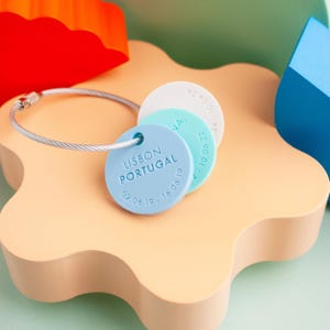 Peut inclure: Un porte-clés bleu avec trois étiquettes rondes. La plus grande étiquette indique "LISBONNE, PORTUGAL 02.06.19 - 16.06.19". Les deux autres étiquettes sont blanches et bleu clair.