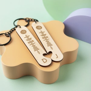 May include: Two wooden keyrings with a heart shape cut out of the bottom. The keyrings are engraved with a sound wave pattern and the letters "D" and "J".