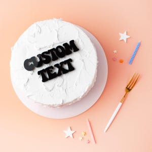 May include: A white cake with black letters that say "CUSTOM TEXT" on top. The cake is on a white plate and there are colourful sprinkles and candles around it.
