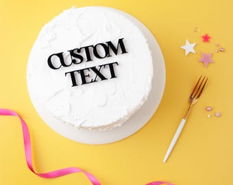 May include: A round white frosted cake with the words "CUSTOM TEXT" in black lettering. A gold and white fork, pink ribbon, and star-shaped decorations are on a yellow background.
