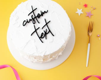 May include: A round cake covered in white frosting with a black acrylic cake topper featuring the words custom text in a cursive script font. The cake is placed on a white circular base, accompanied by a gold fork and small star decorations.