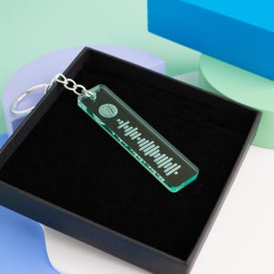 May include: A clear green acrylic keychain with a Spotify logo and a barcode design. The keychain is attached to a silver chain and is displayed in a black box.