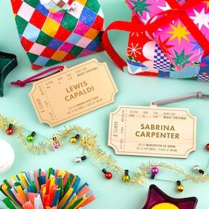 Pu&ograve; includere: Biglietti regalo in legno a forma di biglietti da concerto con i nomi "Lewis Capaldi" e "Sabrina Carpenter". I biglietti includono i dettagli dell'evento e sono circondati da regali confezionati e decorazioni festive.