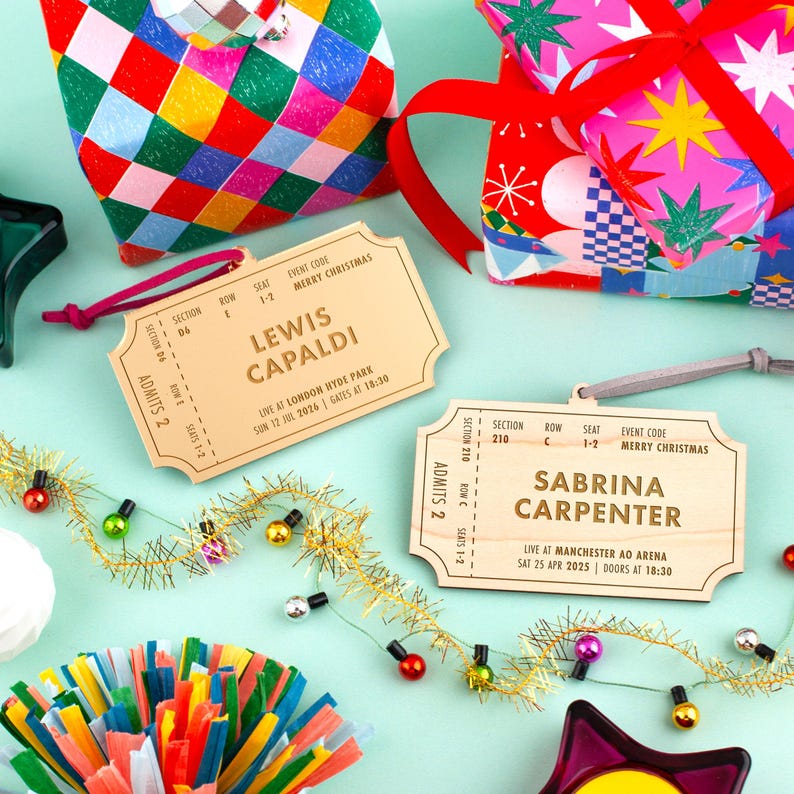 Pu&ograve; includere: Biglietti regalo in legno a forma di biglietti da concerto con i nomi "Lewis Capaldi" e "Sabrina Carpenter". I biglietti includono i dettagli dell'evento e sono circondati da regali confezionati e decorazioni festive.
