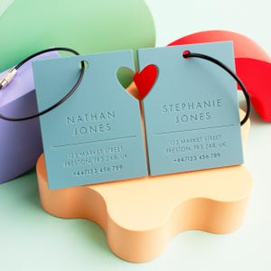May include: Two blue luggage tags with a heart-shaped cutout. The tags are connected by a black cord. The text on the tags reads "Nathan Jones" and "Stephanie Jones" with their respective addresses and phone numbers.