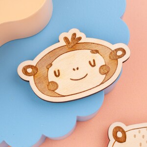 May include: A wooden monkey-shaped craft piece with a smiling face, brown details, and a light wood finish. The monkey is set against a blue, scalloped background, with a pink surface visible.